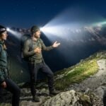 Tres senderistas en la oscuridad total de la montaña, cada uno activando una linterna frontal diferente (Petzl, Ledlenser y Black Diamond), mostrando la diferencia real en el alcance y apertura de los haces de luz.